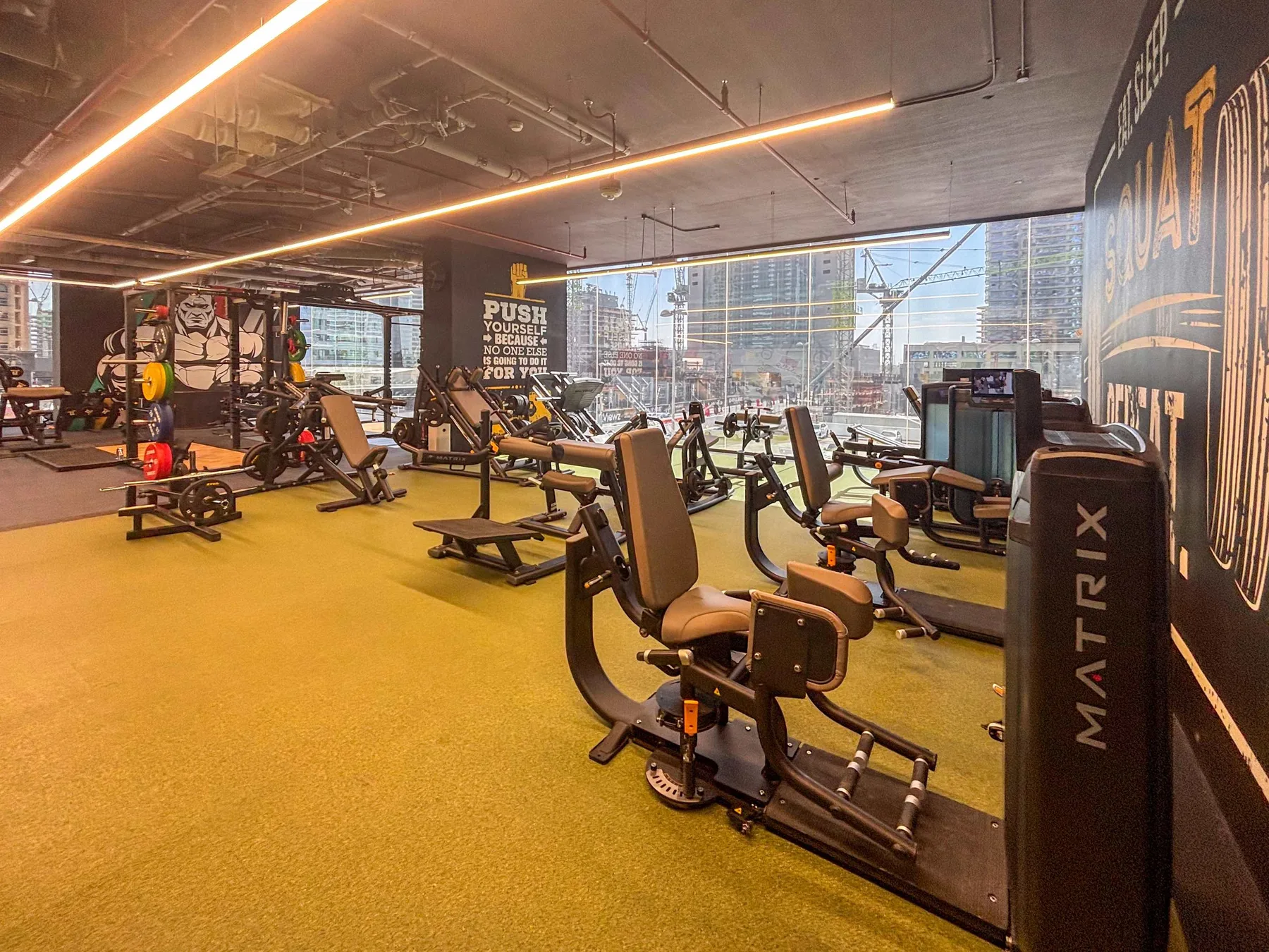 Un gimnasio con máquinas de ejercicio y grandes ventanas con vistas a edificios de la ciudad.