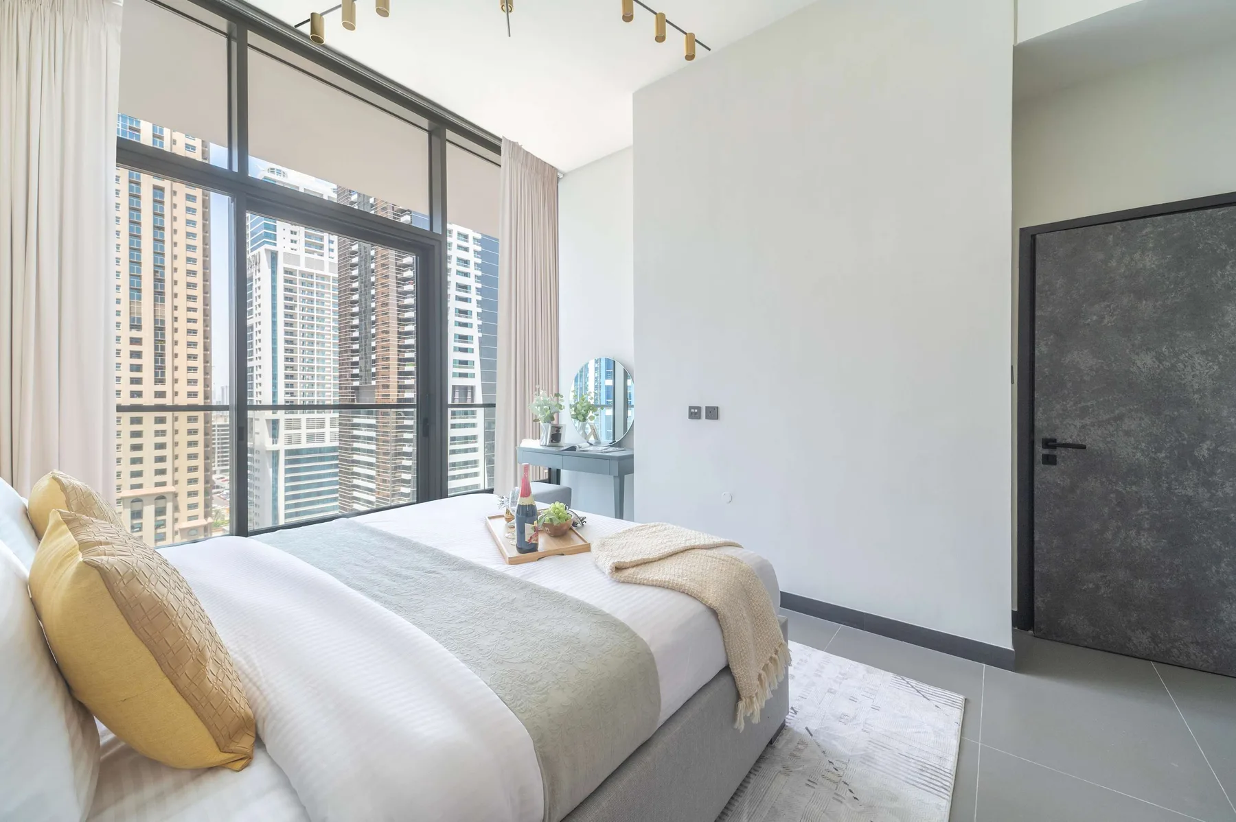 Un dormitorio luminoso con una cama junto a grandes ventanas con vistas a edificios altos.