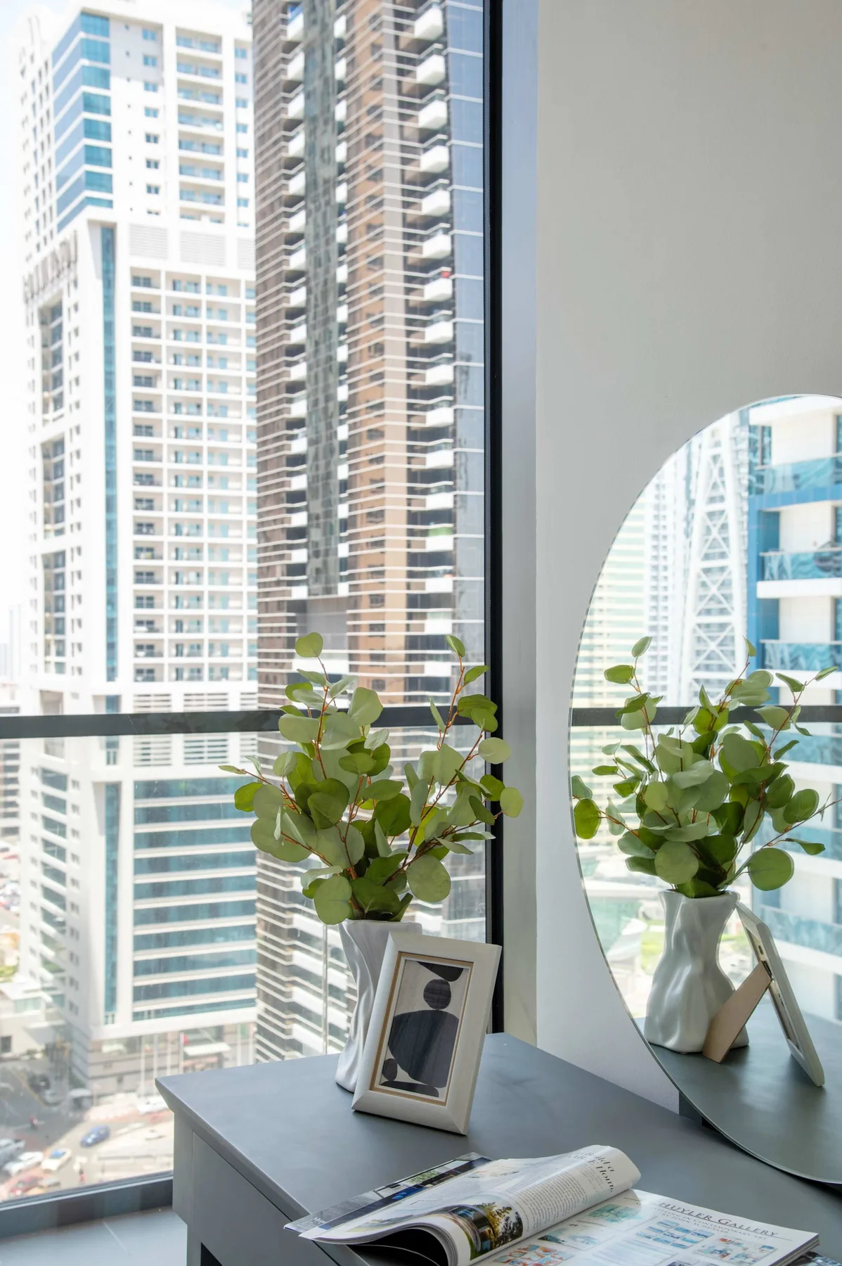 Una habitación luminosa con una gran ventana, edificios de la ciudad y una planta frondosa sobre una mesa.