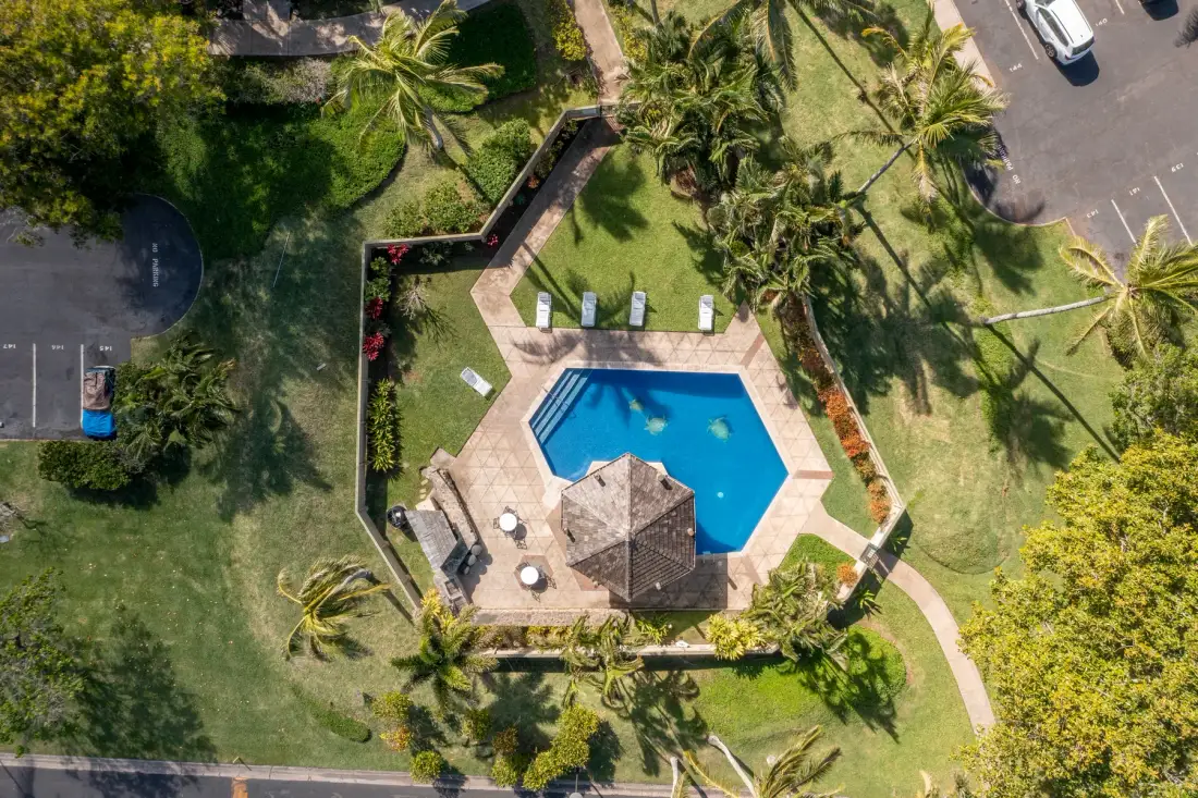 An aerial view shows a swimming pool surrounded by a paved patio and greenery.