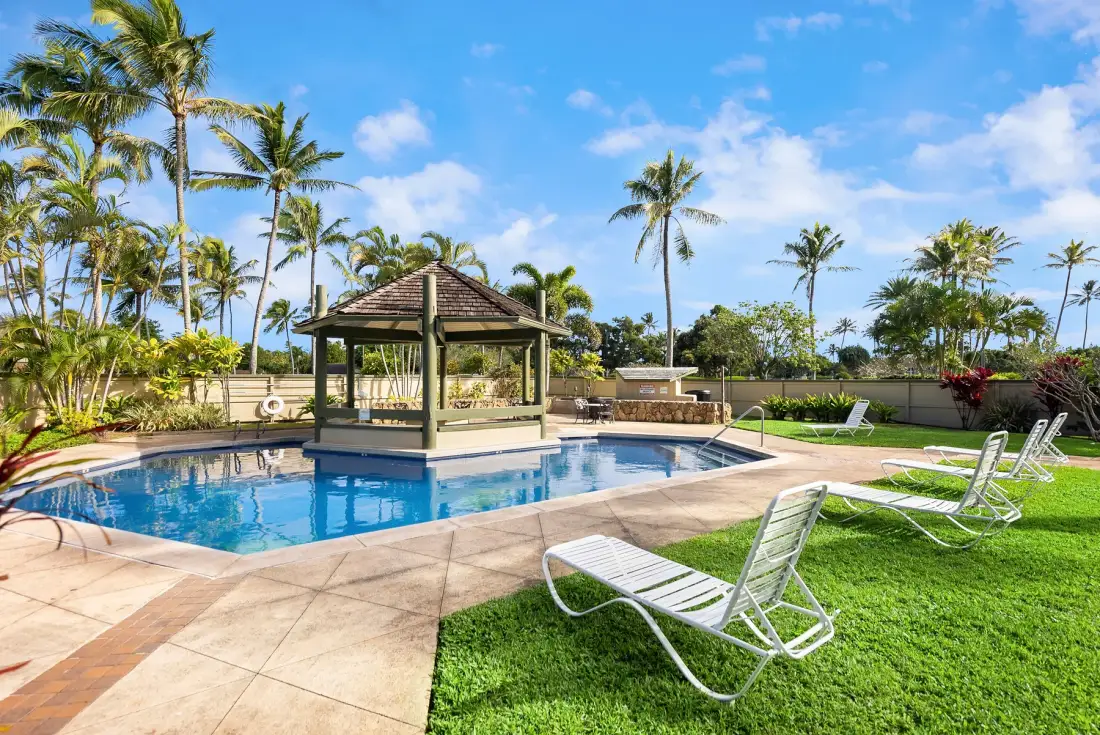 An outdoor pool area with a shaded pavilion, palm trees, and lounge chairs.