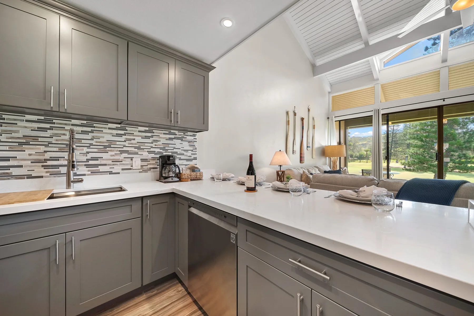A bright kitchen with a long counter overlooking a living area and large windows.