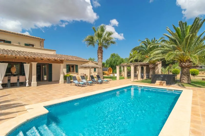 Una piscina junto a un patio de baldosas, con una casa y palmeras cerca.
