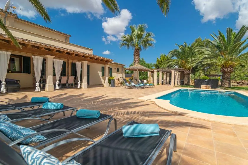Un patio junto a una piscina, con asientos reclinables y palmeras.