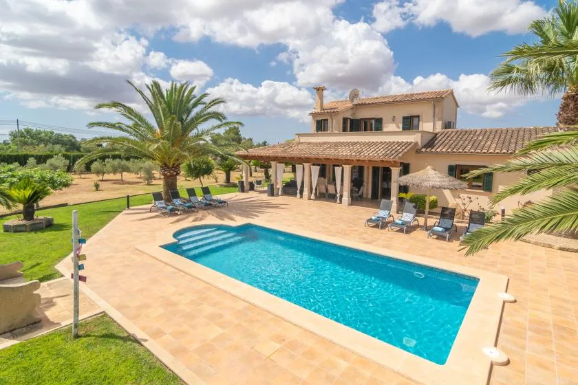 Un patio junto a una piscina y una casa grande, con asientos y palmeras.