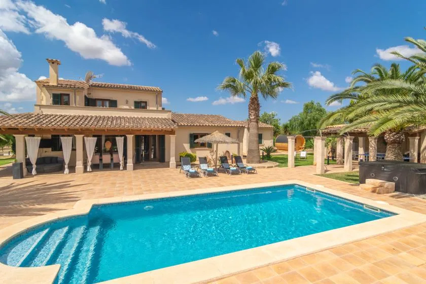 Una piscina junto a un patio de baldosas, tumbonas y una villa.