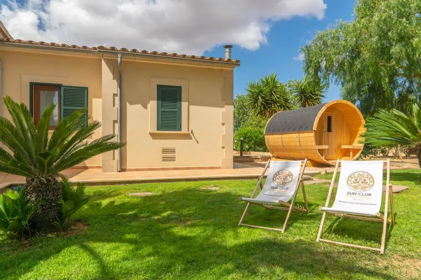 Un jardín soleado junto a una casa, con dos tumbonas y una estructura cilíndrica de madera.