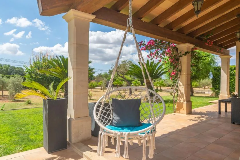Un patio cubierto con una silla colgante da a un jardín verde.