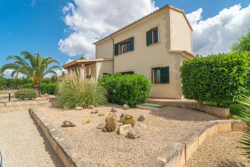 Una casa beige de dos plantas junto a un jardín de grava con arbustos y una palmera.