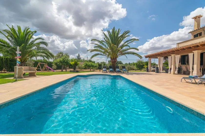 Una piscina junto a un patio pavimentado y palmeras bajo un cielo parcialmente nublado.
