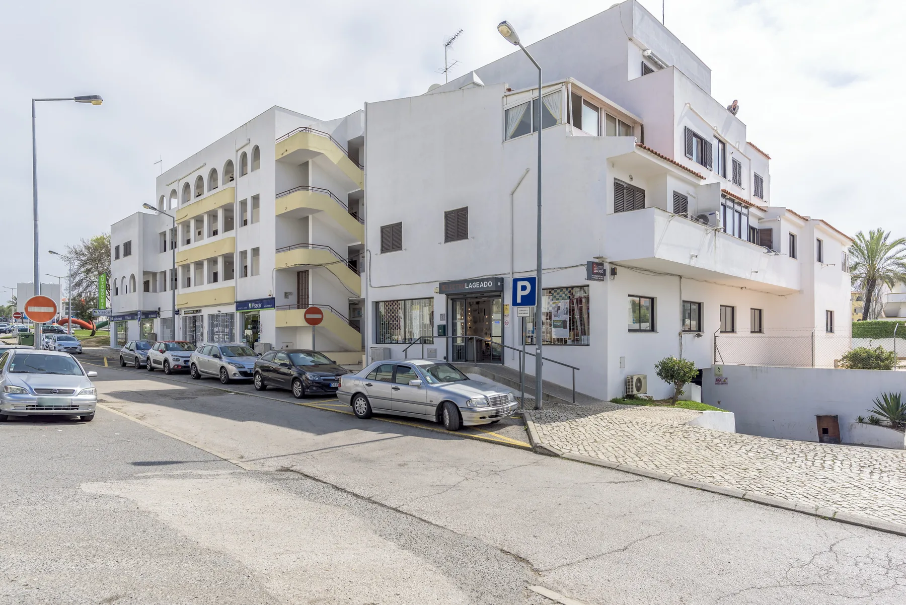 A street scene with white apartment buildings, parked cars, and a sloping roadway.