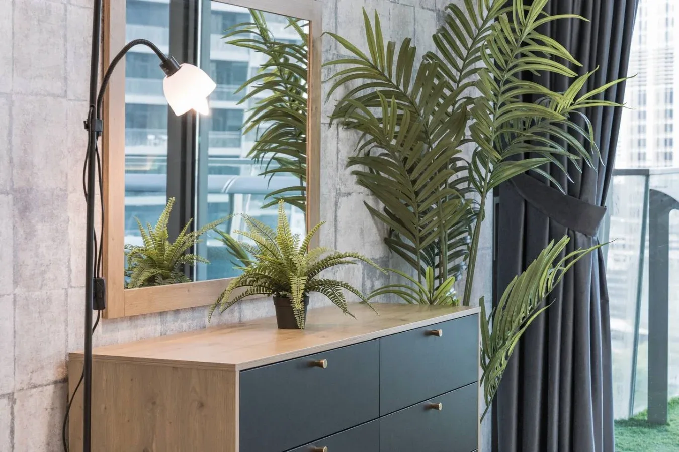 Un rincón de dormitorio con una cómoda, plantas frondosas y una lámpara de pie junto a grandes ventanas.