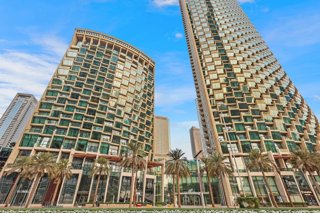 Edificios modernos de gran altura con ventanas de vidrio se alzan sobre palmeras bajo un cielo azul.
