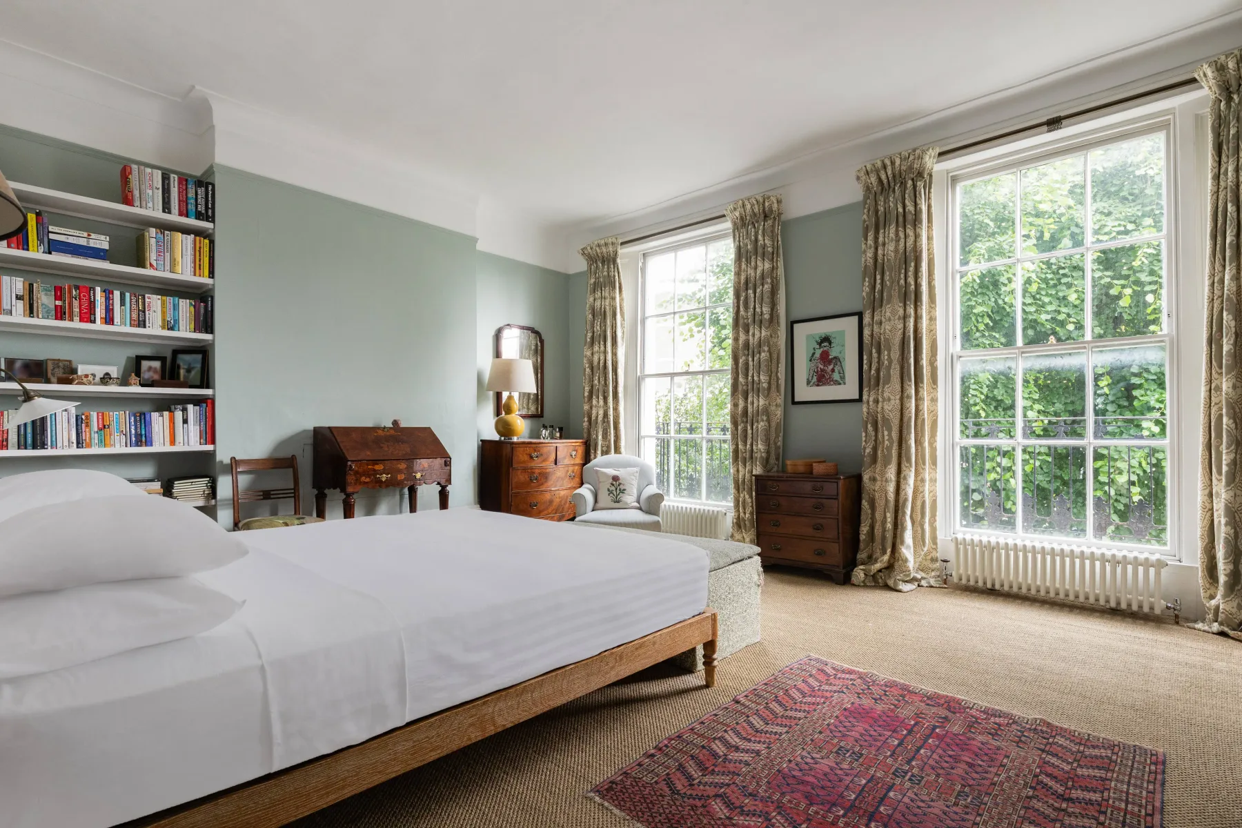 A bedroom with a bed, bookshelves, and tall windows.