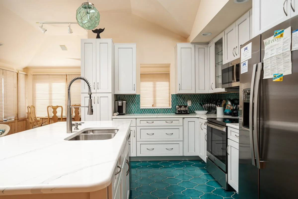 Una cocina luminosa con gabinetes blancos, una isla central y azulejos verde azulado.