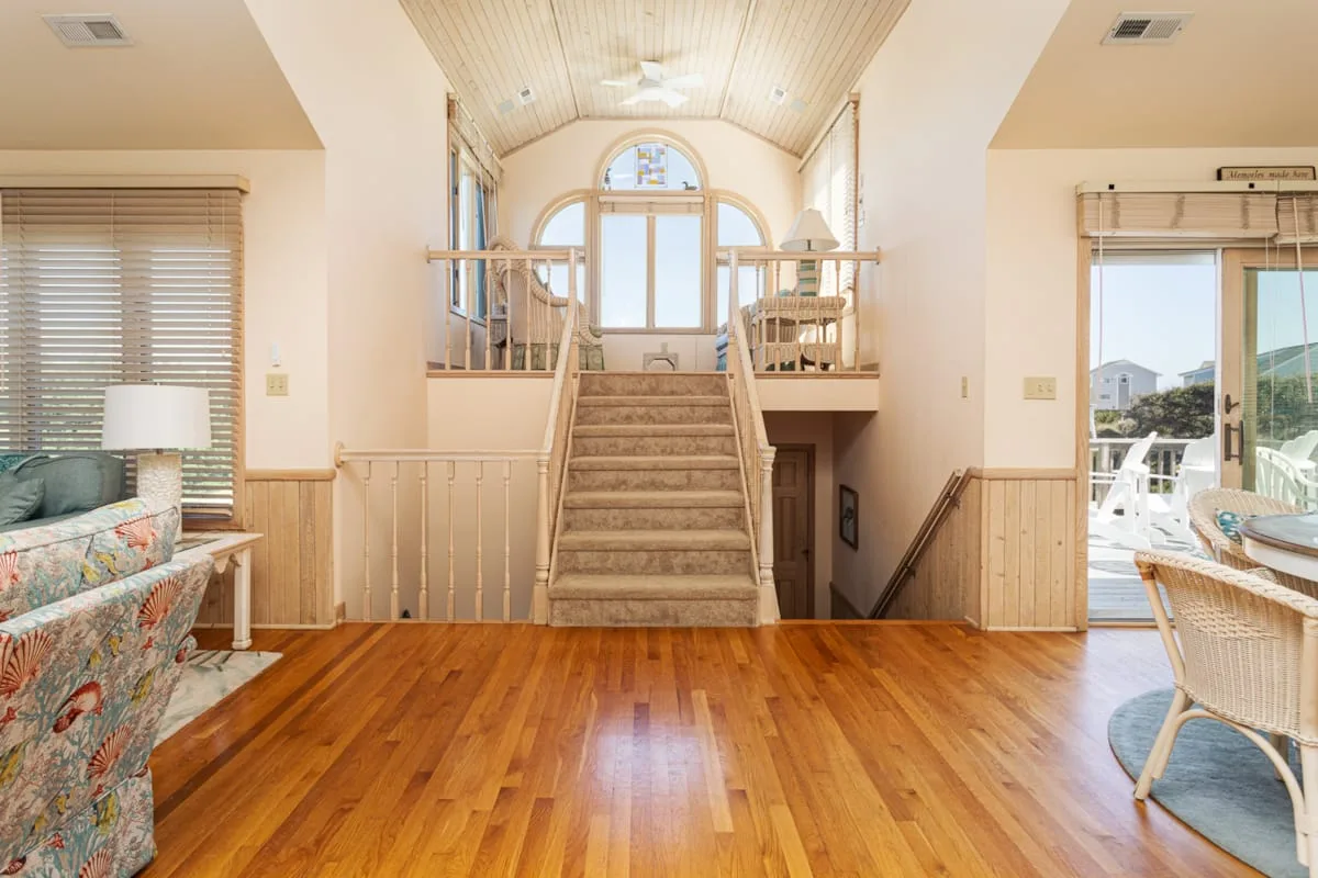 Una sala luminosa se abre a una escalera y un rellano superior soleado.