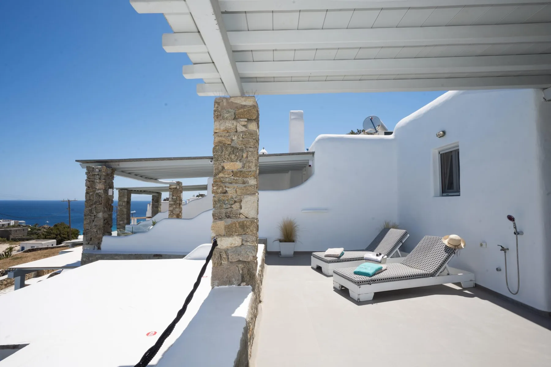 A sunlit rooftop terrace with white walls, stone columns, and lounge seating.