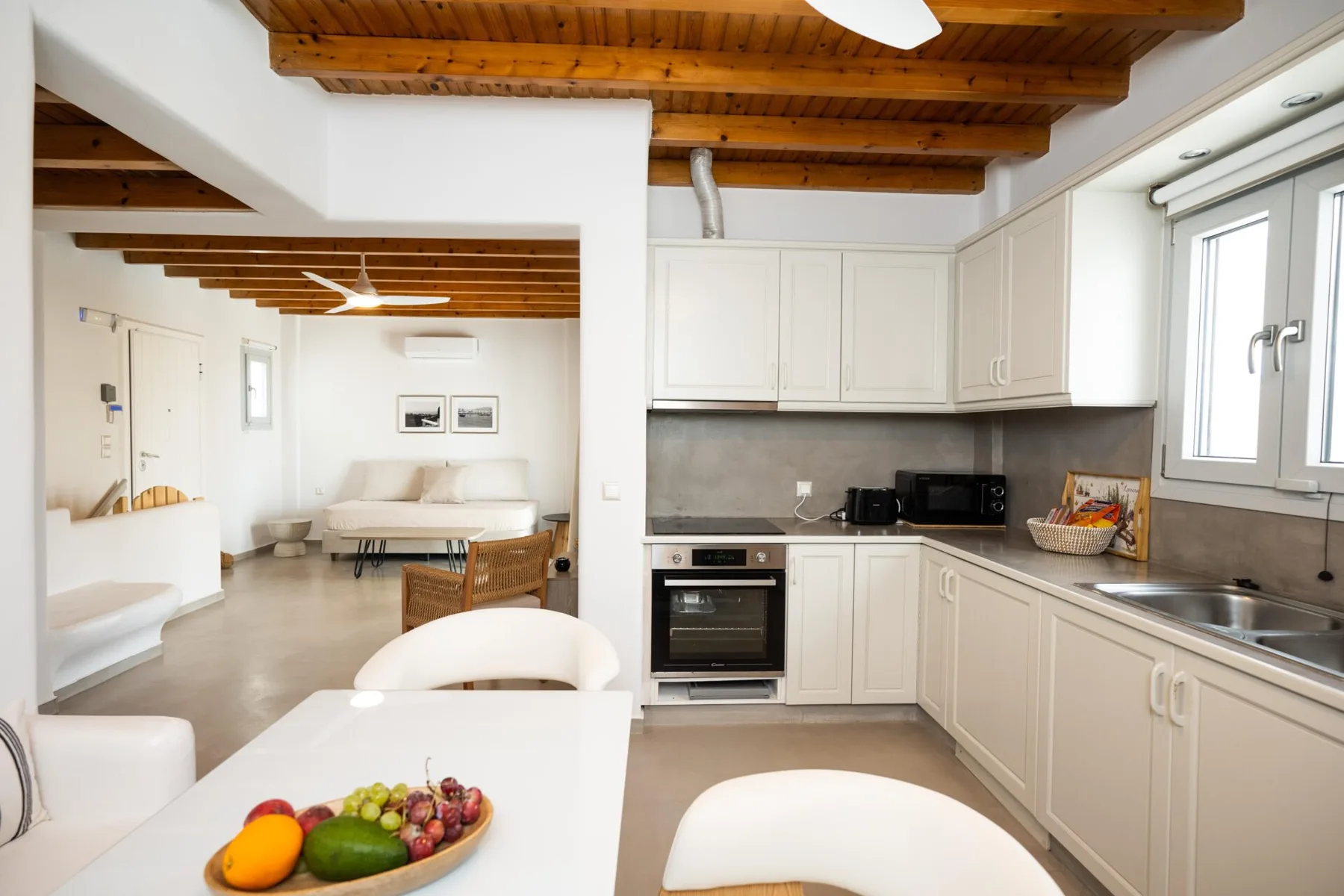 A bright open-plan kitchen and dining area with white cabinets and exposed wooden ceiling beams.