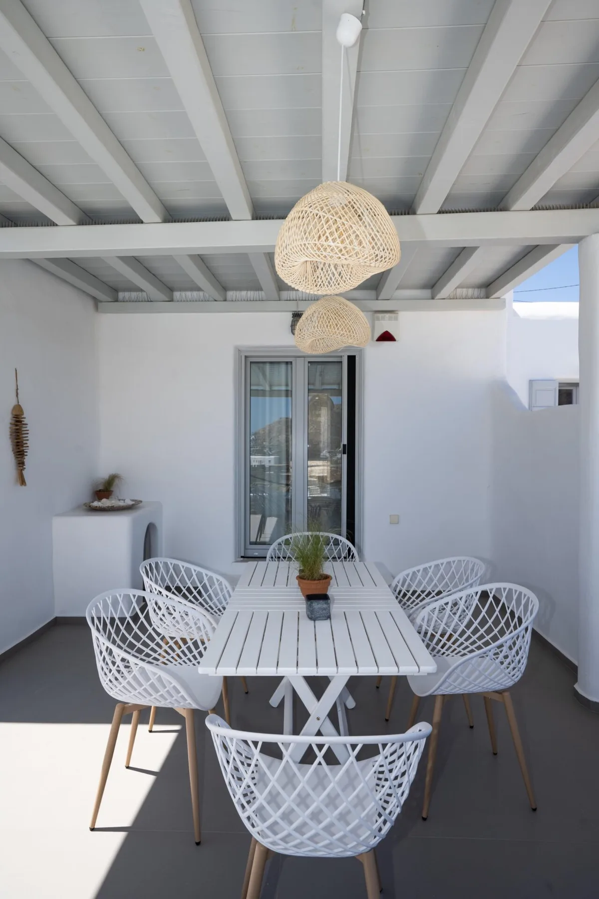 An outdoor dining area has a long table and white woven seating beneath pendant lights.