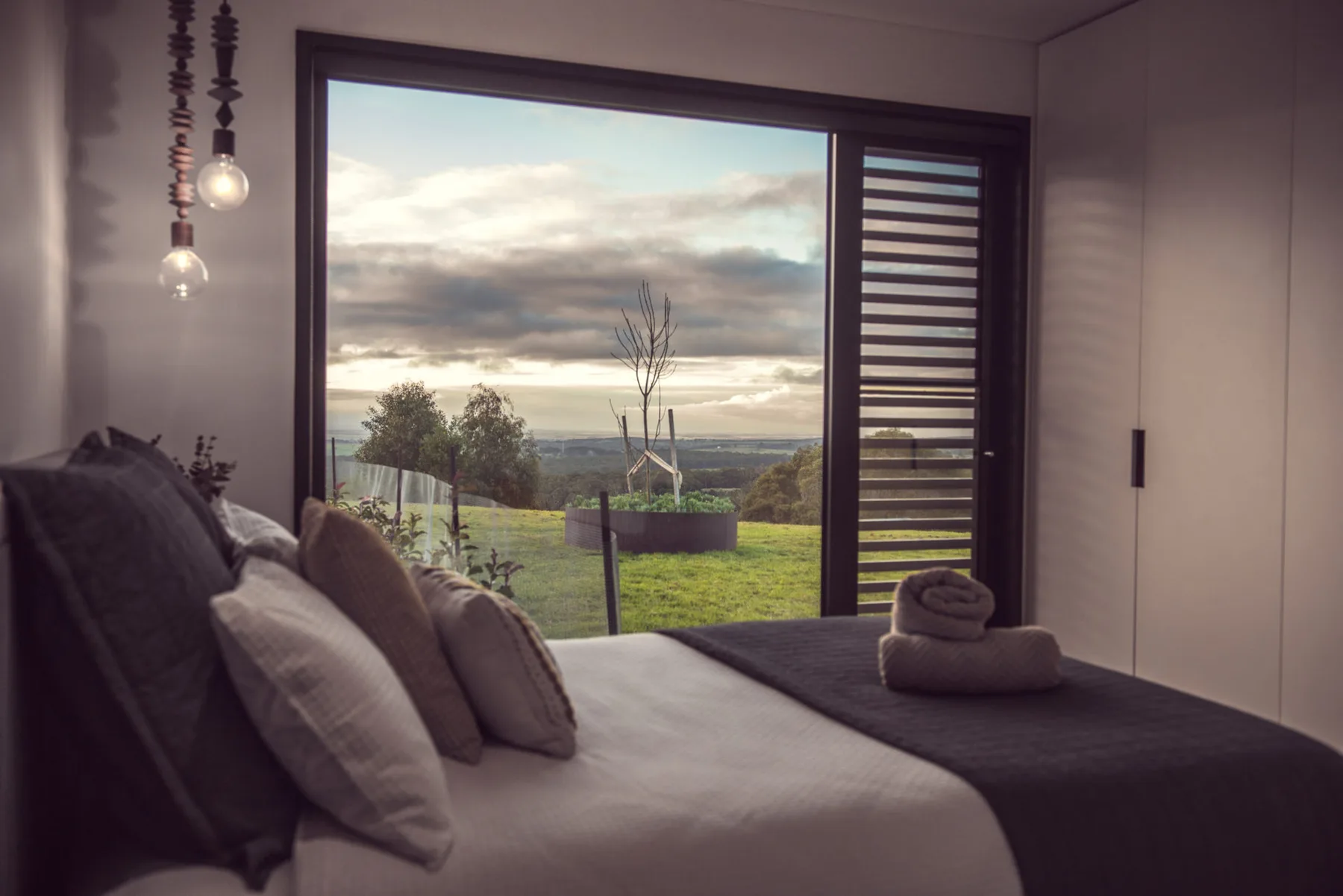A bedroom with a neatly made bed facing a large window overlooking a green landscape.