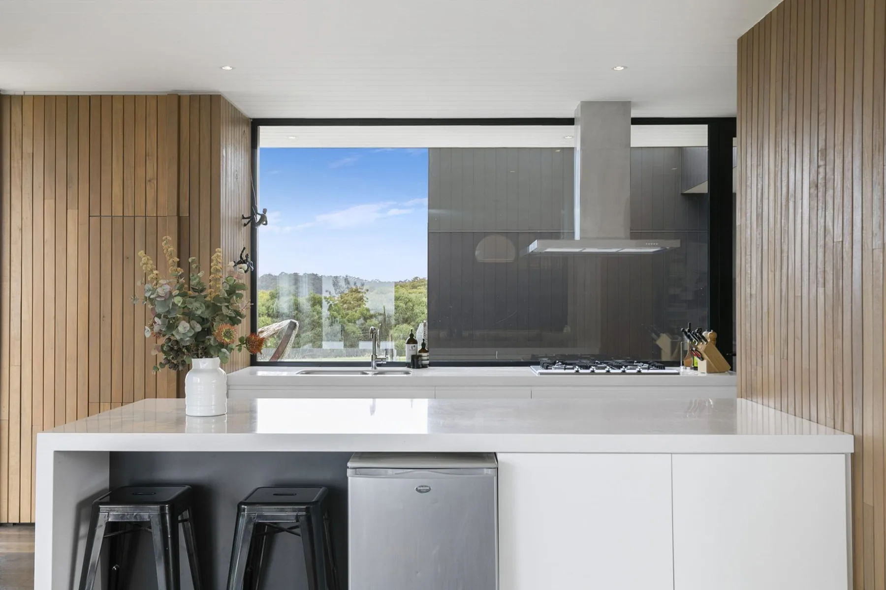 A modern kitchen with a white island and a wide window overlooking greenery.