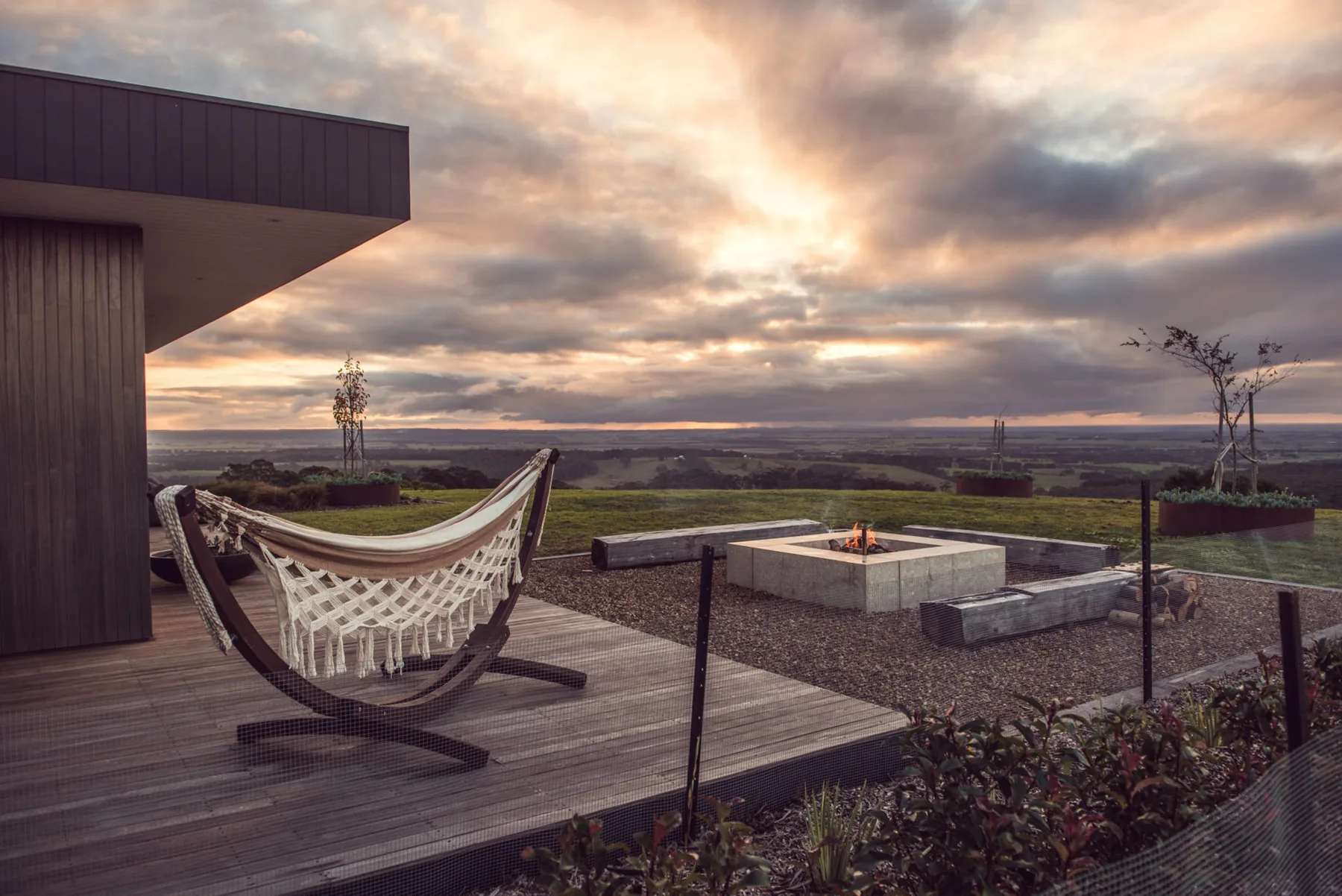 A hammock rests on a wooden deck beside a low outdoor fire pit at sunset.