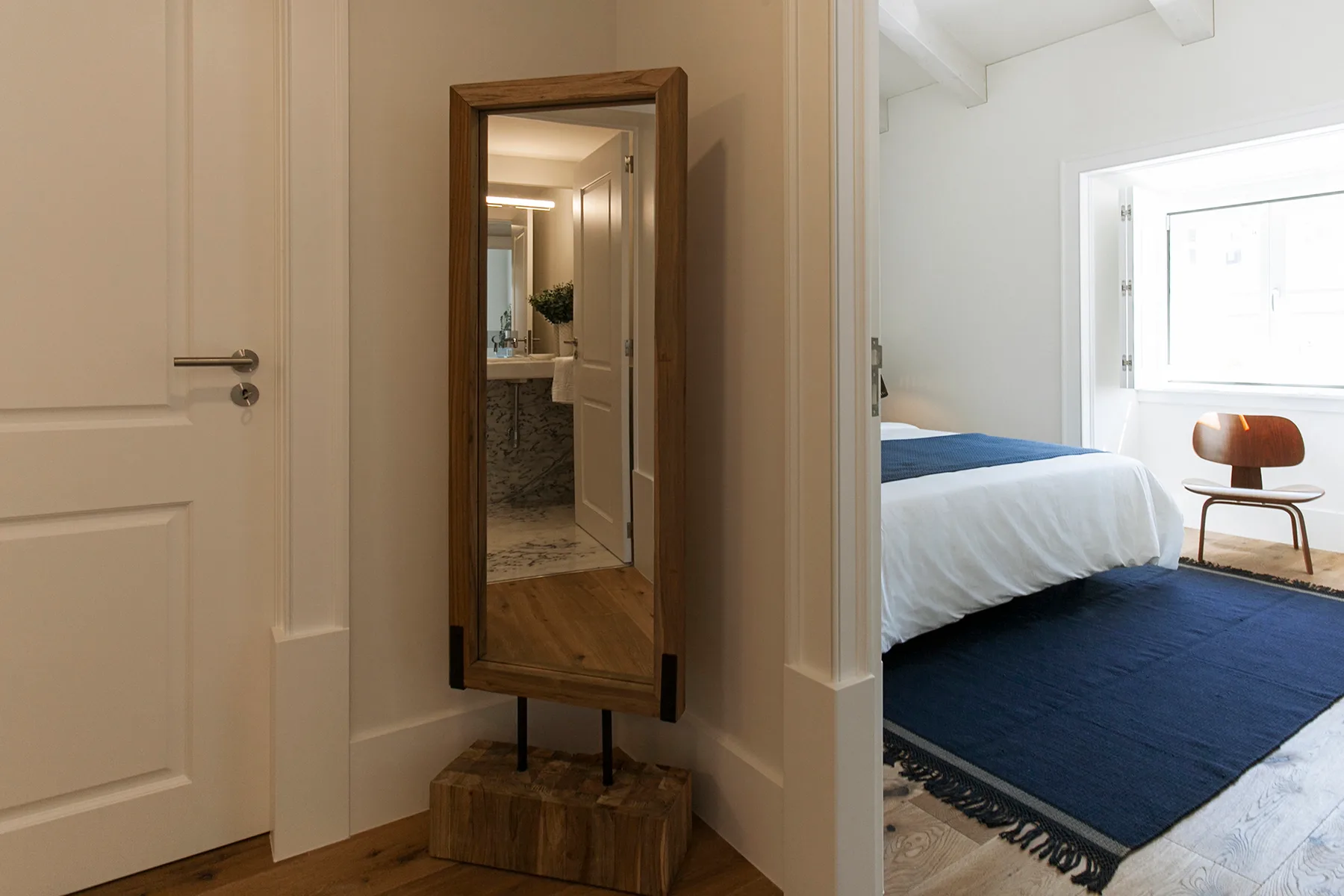 A bright bedroom with a bed, blue rug, and chair beside the window.