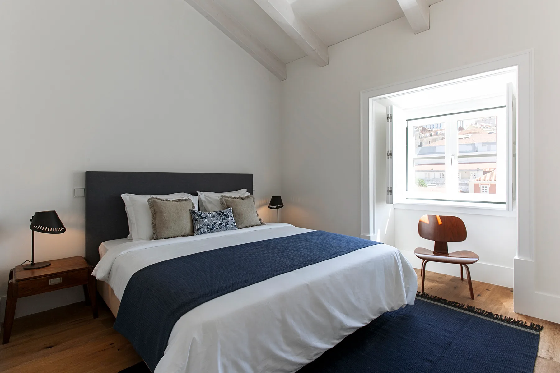 A bright bedroom with a neatly made bed and a window overlooking nearby buildings.