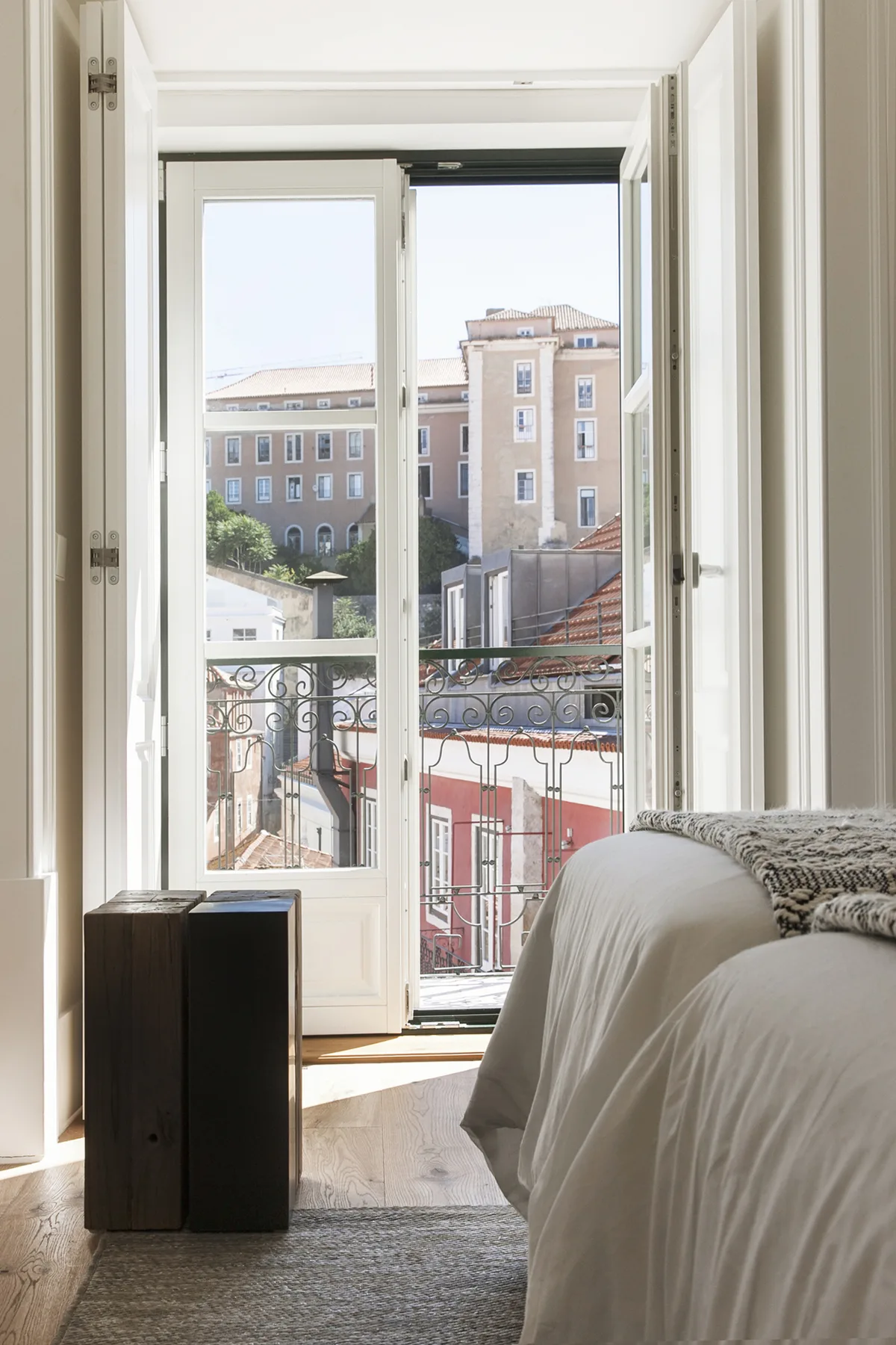 A bright bedroom with a bed beside an open window overlooking nearby buildings.