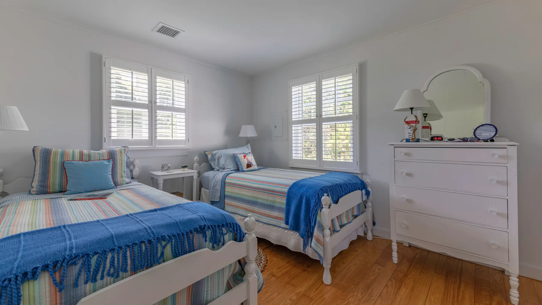 A bright bedroom with two beds, colorful striped bedding, and white shutters.