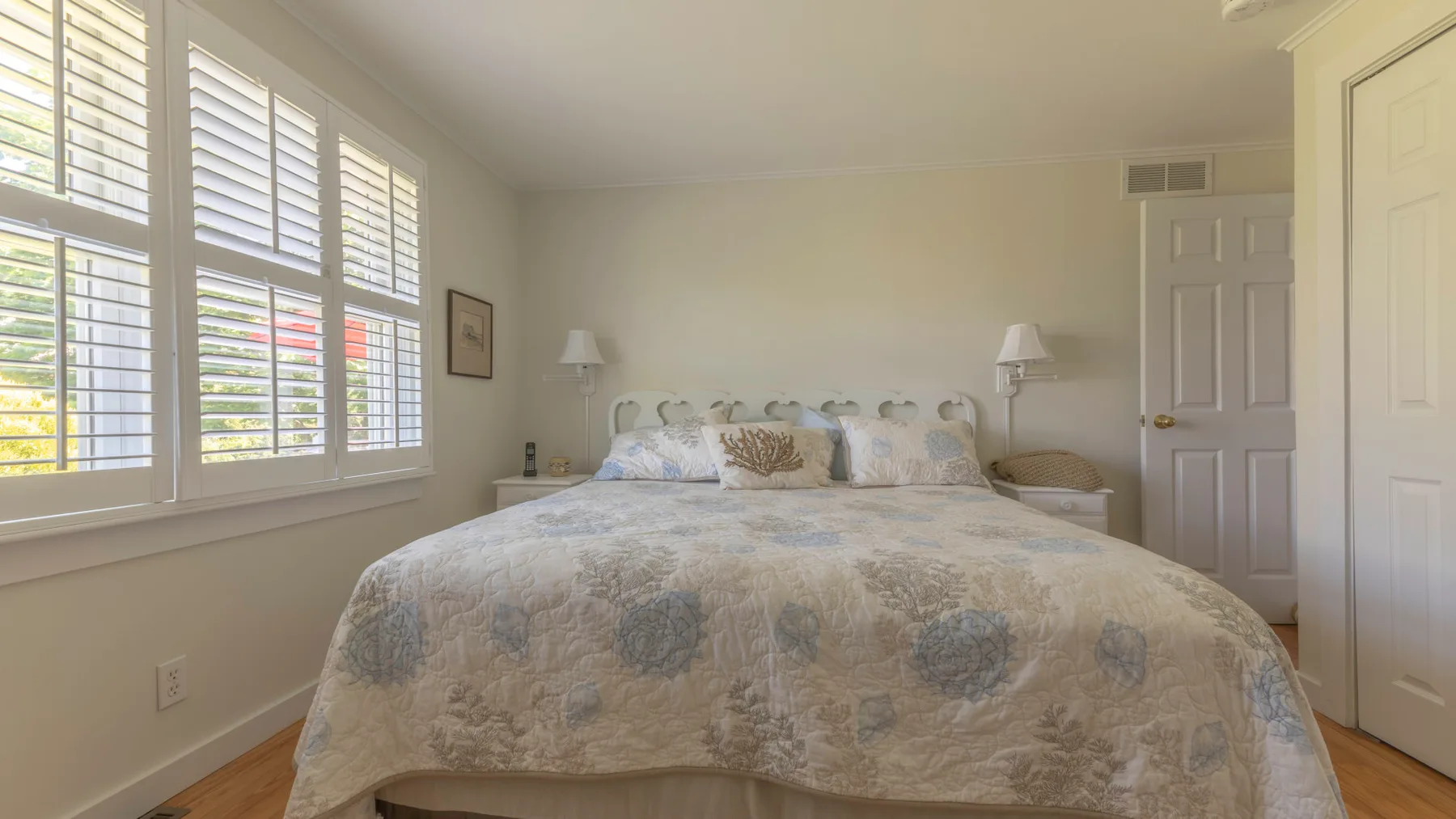 A bedroom with a large bed beneath several windows with white shutters.