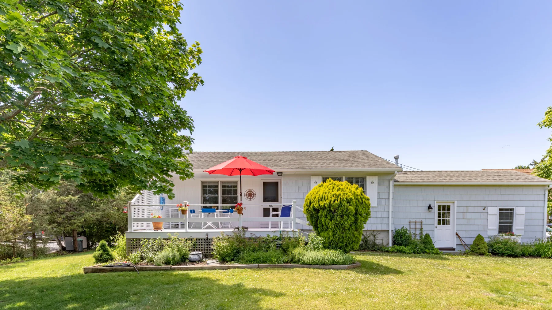 A single-story house stands behind a lawn, with a deck and leafy trees.