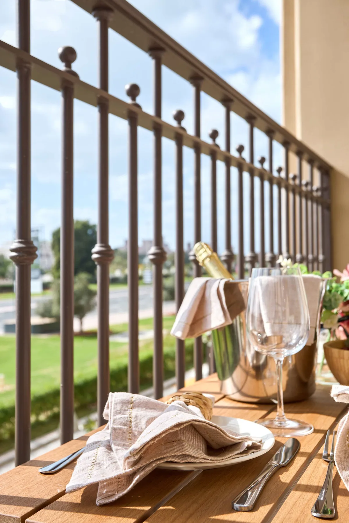Una mesa de comedor al aire libre, iluminada por el sol, está junto a una barandilla metálica con vistas a zonas verdes.