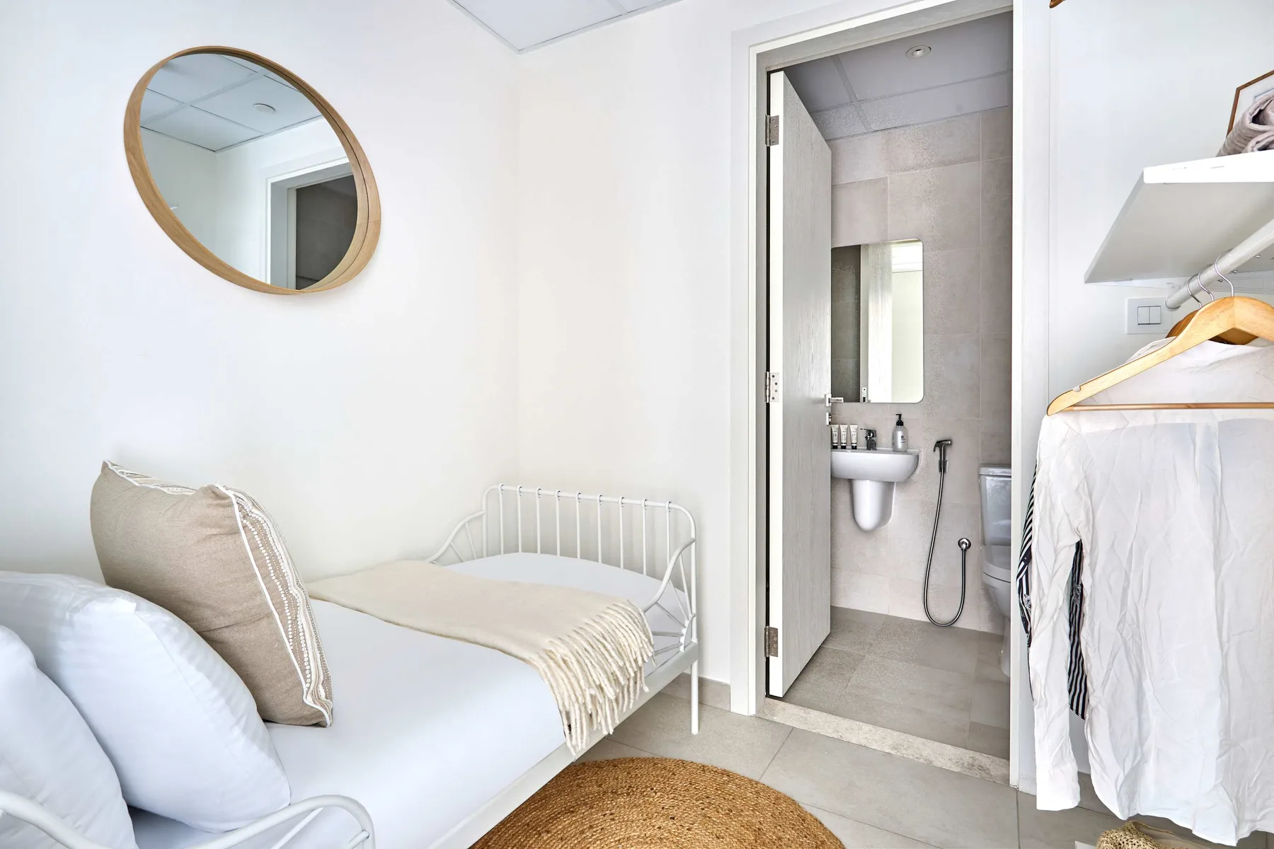 Un dormitorio luminoso con una cama, un espejo redondo y la puerta abierta al baño.