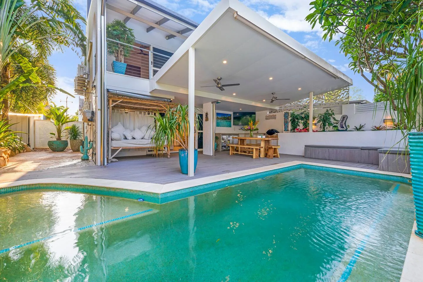 An open-sided patio overlooks a swimming pool, with seating and tropical plants nearby.