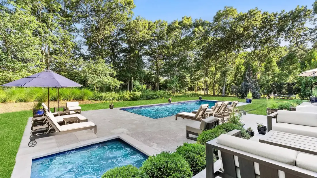Una piscina al aire libre y un patio con asientos, vegetación y árboles alrededor.