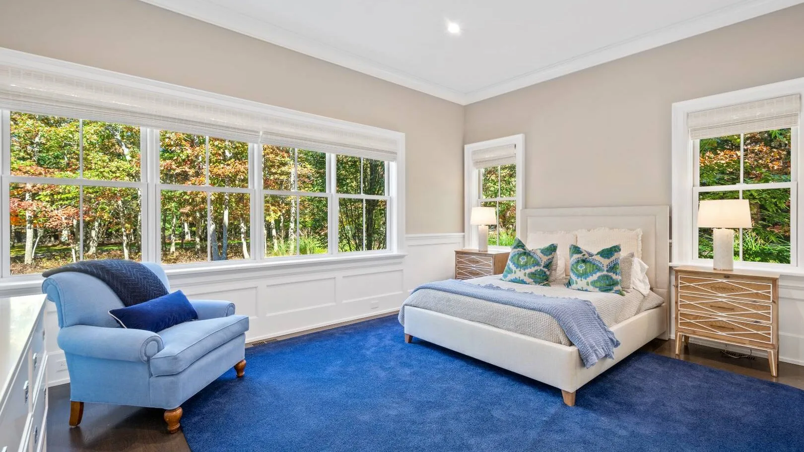 Un dormitorio luminoso con una cama, un sillón azul y grandes ventanas con vista a los árboles.