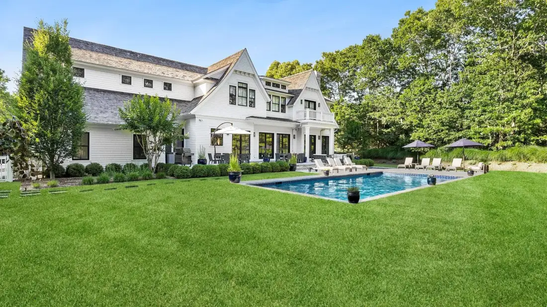 Una gran casa blanca está junto a una piscina y un amplio césped verde.