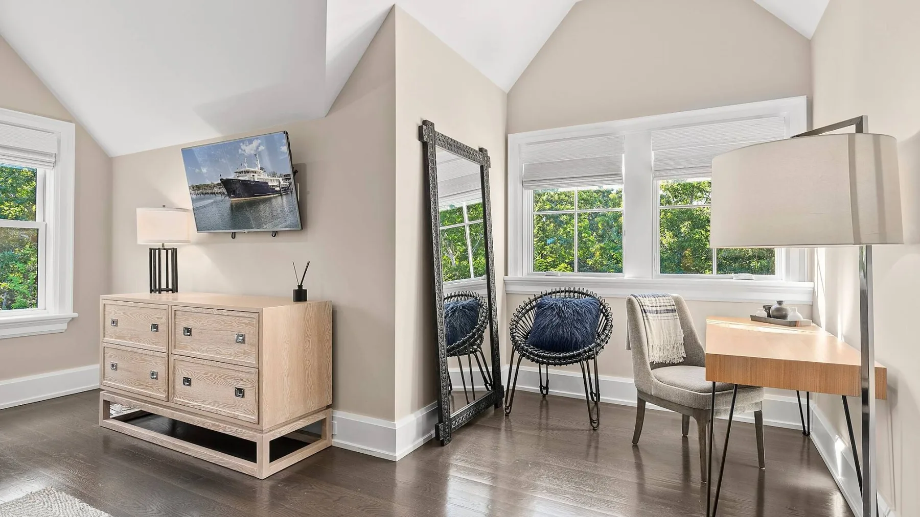 Una habitación luminosa tiene una cómoda con un televisor montado y una mesa pequeña junto a las ventanas.