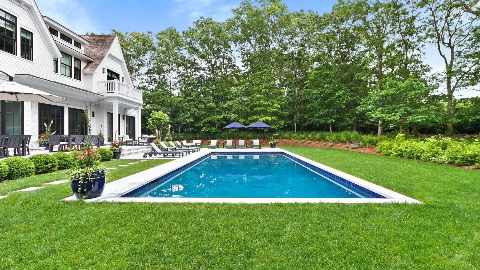 Una piscina en el patio trasero está junto a una casa blanca y un césped verde.
