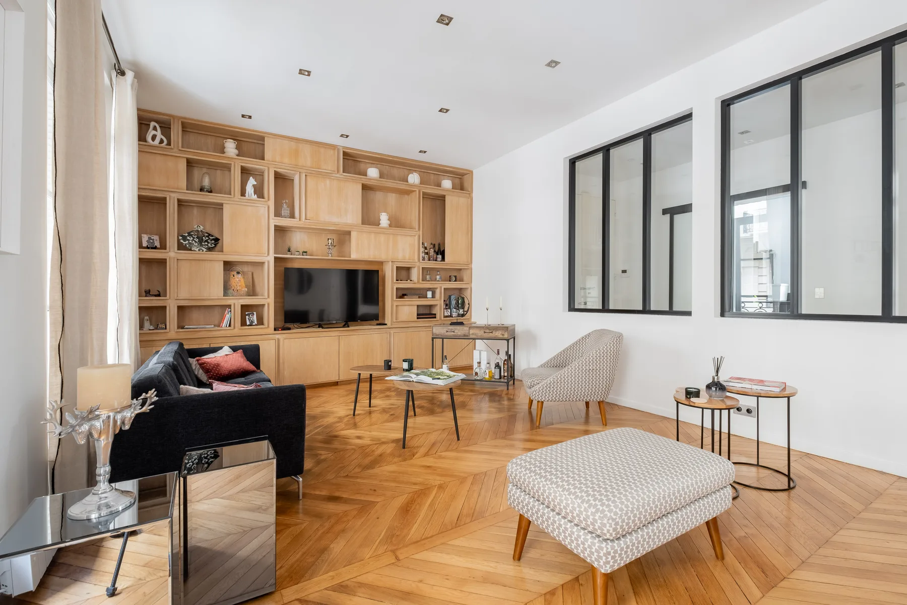 Una sala de estar luminosa con estanterías de madera, asientos y grandes ventanas con marcos negros.