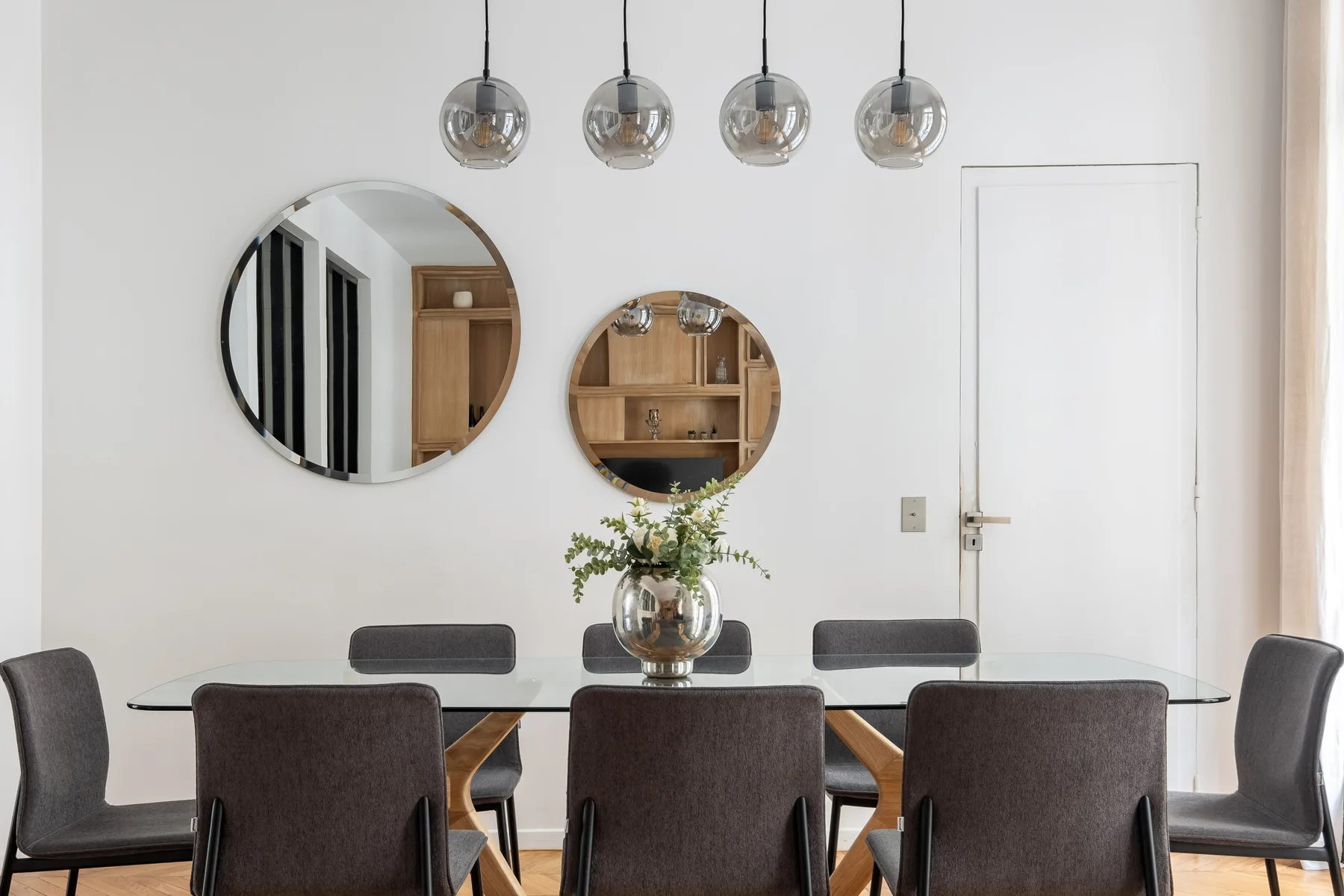 Comedor con mesa, asientos tapizados, lámparas colgantes y espejos redondos en la pared.