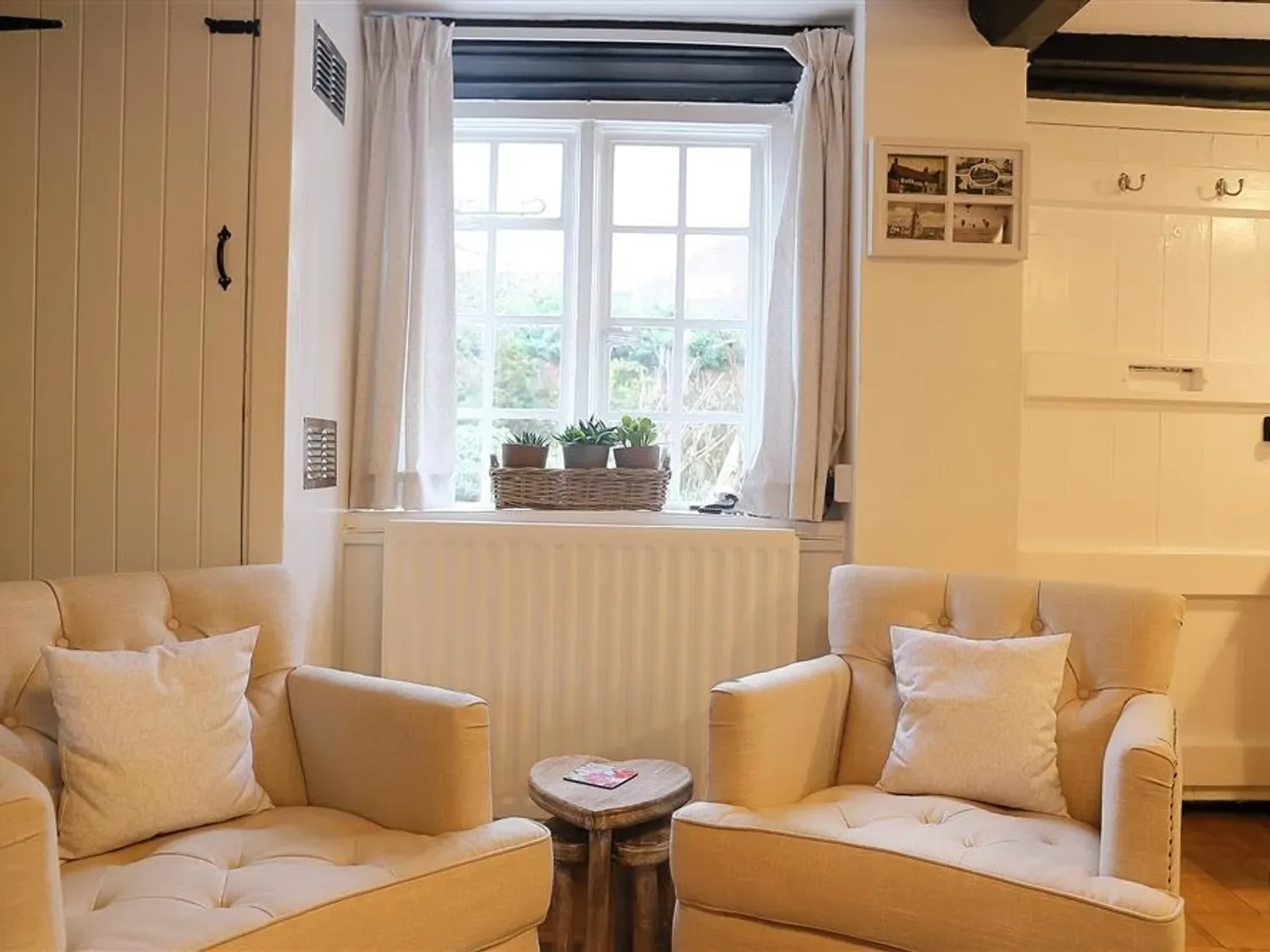 A sitting area with two beige armchairs a small wooden table between them and a window with potted plants at One Masons Court in Stratford upon Avon