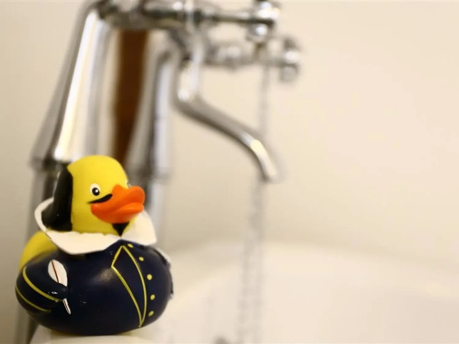 A rubber duck wearing a dark costume sits beside a faucet in a bathroom sink area.