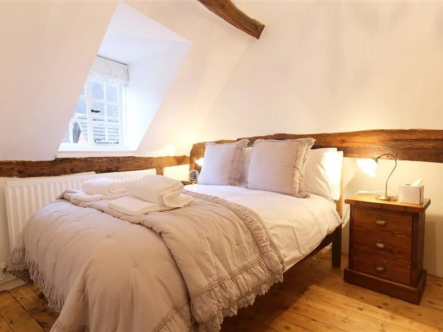 A bedroom with a double bed with beige bedding and pillows next to a wooden nightstand with a lamp at One Masons Court in Stratford upon Avon