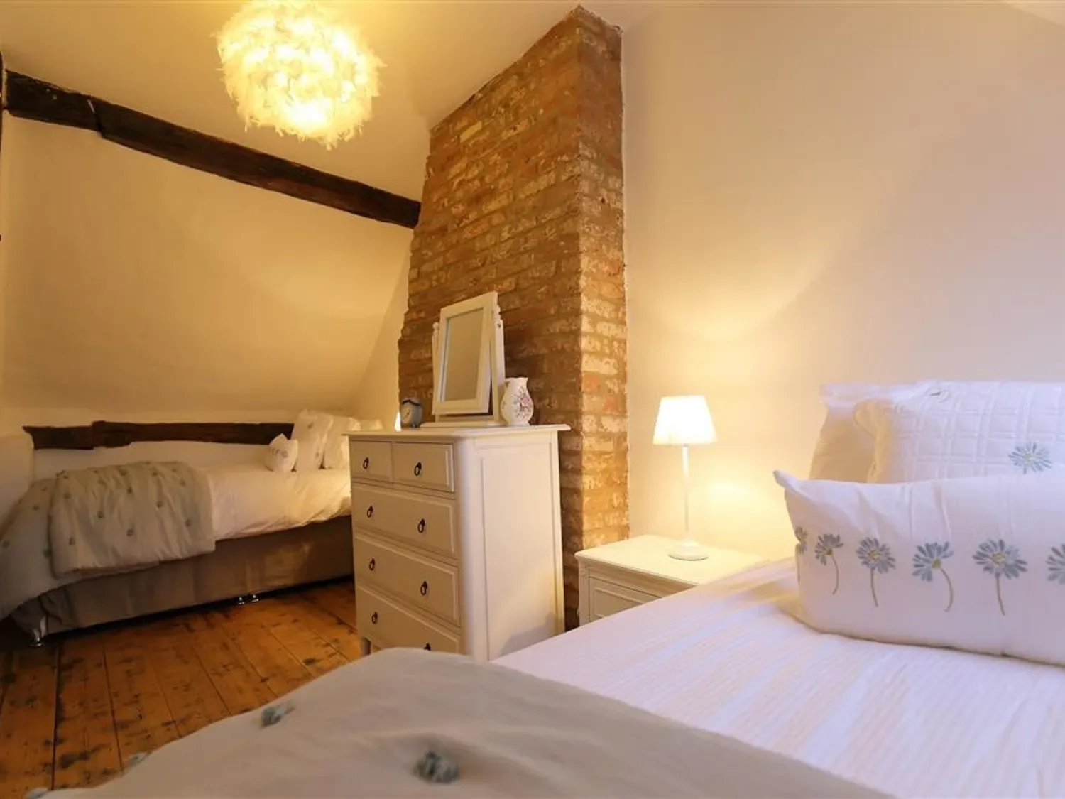 A bedroom with two beds a chest of drawers a bedside table with a lamp and exposed brick wall at One Masons Court in Stratford upon Avon