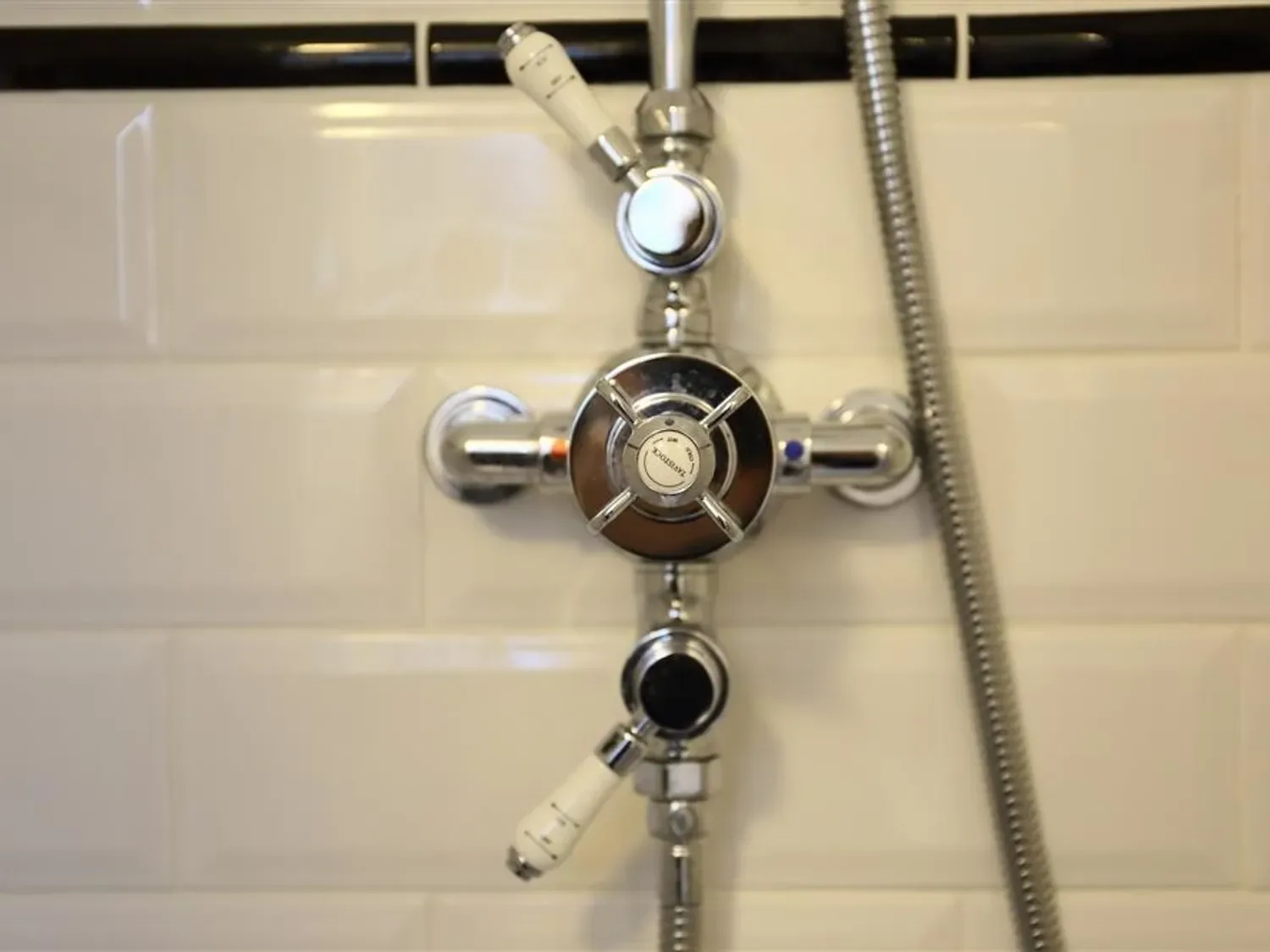 A close up of a shower control valve with white handles and a flexible metal hose attached on a tiled wall at One Masons Court in Stratford upon Avon