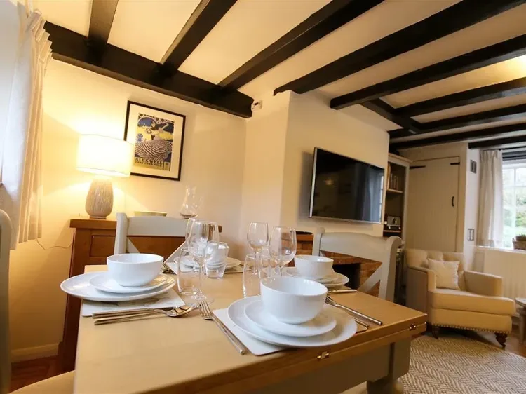 A dining table set with dishes and glasses sits in a living room with exposed ceiling beams.