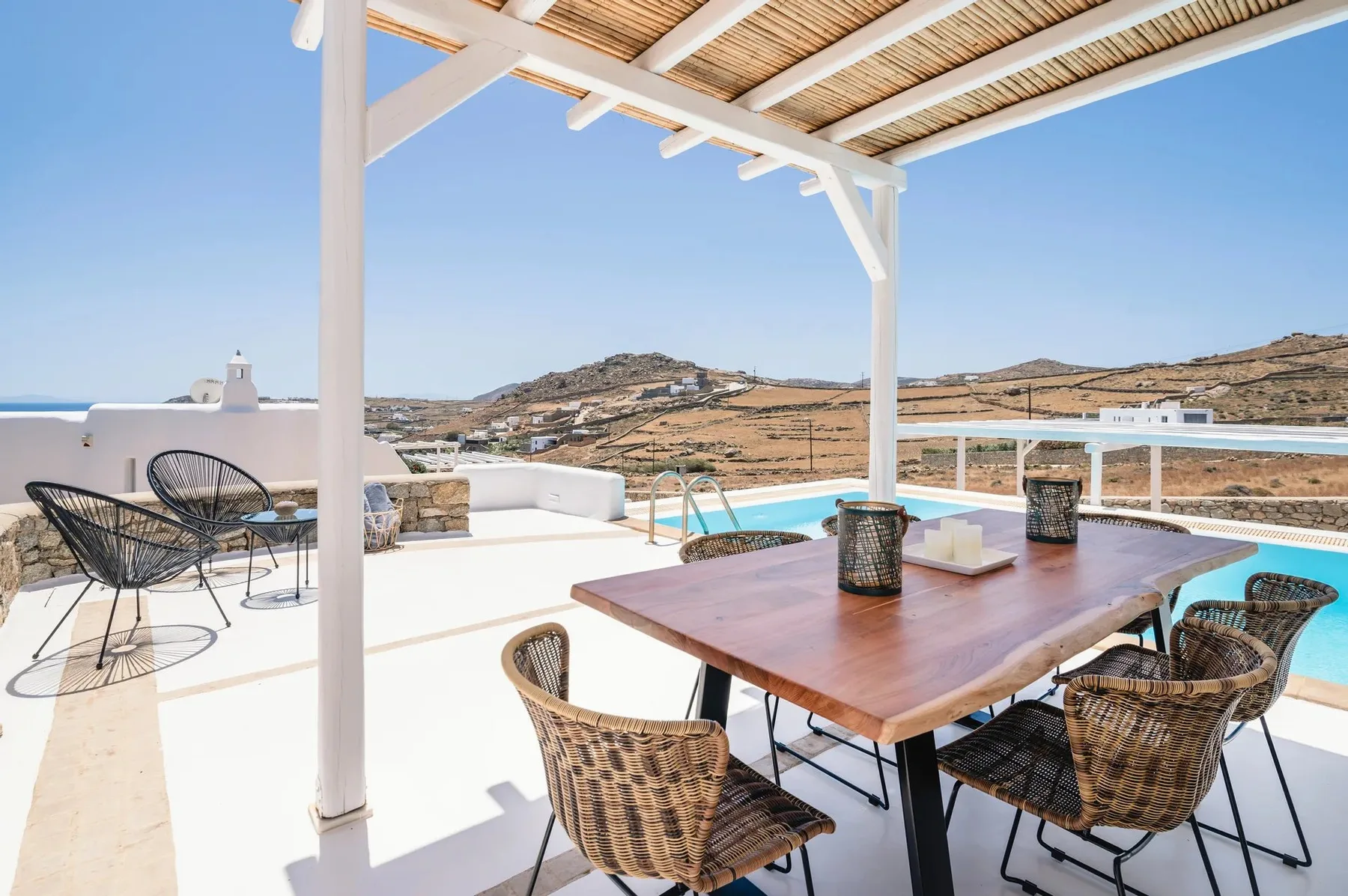 Terraza exterior con mesa de madera, asientos tejidos y vistas a las colinas.