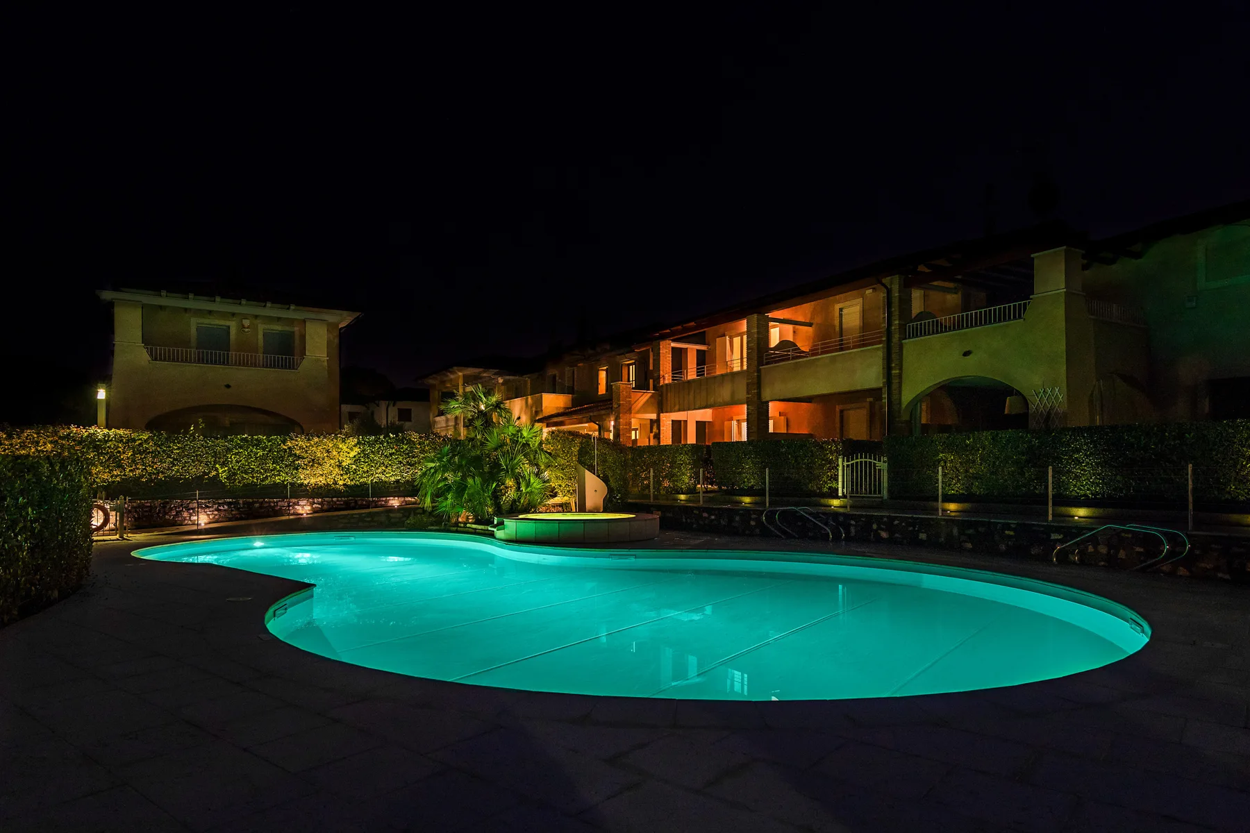 A brightly lit swimming pool sits in front of a row of buildings at night.